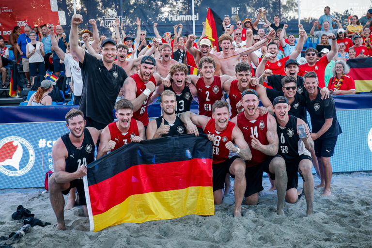 A group of beach volleyball players and staff celebrate on the sand, holding a German flag, with cheering fans in the background.