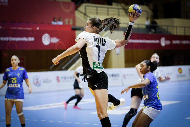 An athlete in a white and black uniform leaps to pass a handball during an indoor match, with opponents in blue uniforms nearby.