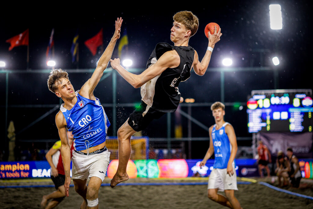 Two athletes leap in mid-air during a beach handball match, one holding the ball while the other reaches up to block.
