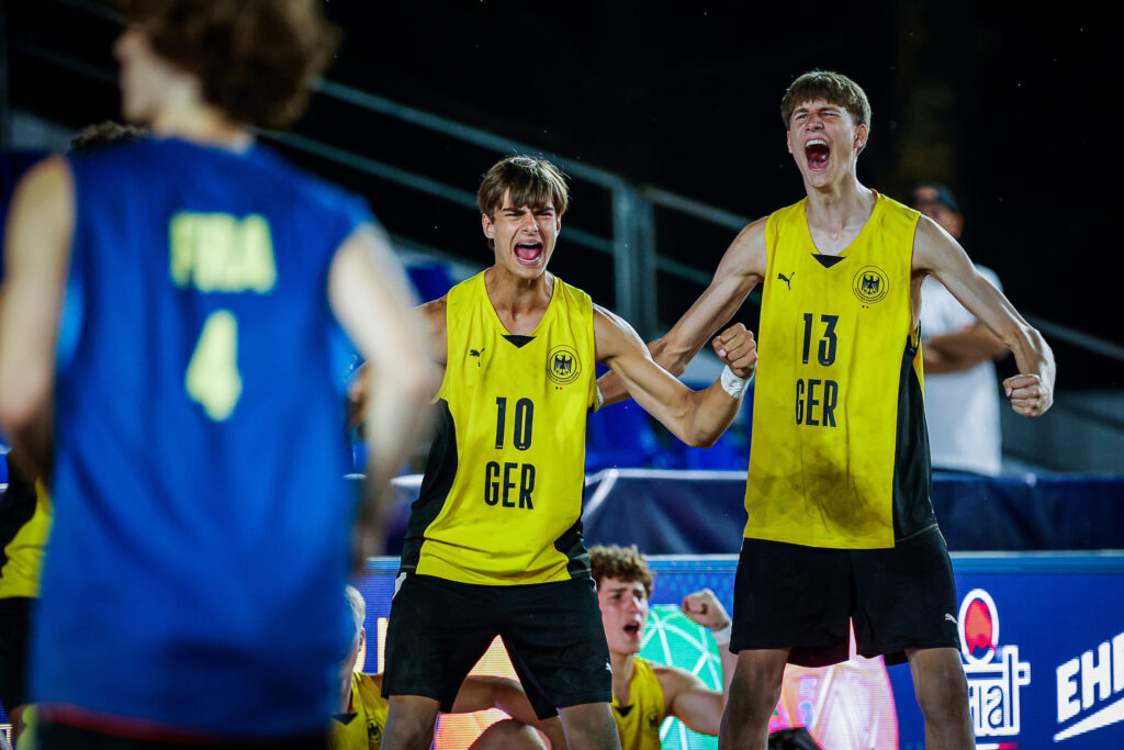 Two athletes in yellow jerseys with "GER" and numbers 10 and 13 celebrate enthusiastically on a court, while an opponent in a blue jersey is seen from behind.