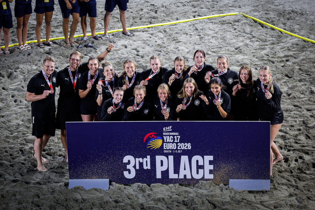 A team of athletes stands on a beach holding medals and posing behind a sign that reads “3rd Place” for the EHF Beach Handball Euro 2026.