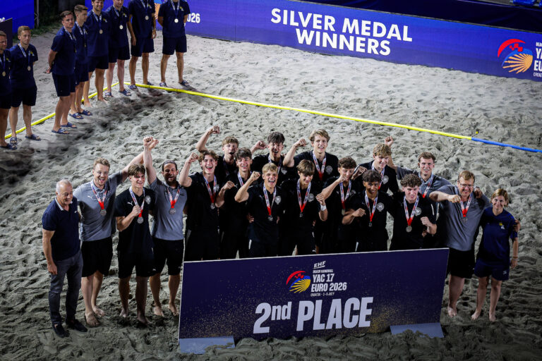A group of athletes and staff celebrate on a sandy beach, wearing medals and posing with a sign that reads "2nd PLACE" at the EHE Beach Handball Euro 2026.
