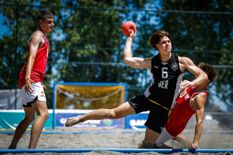 An athlete in a black jersey with the number 6 leaps to throw a ball while being challenged by an opponent on a sandy court.