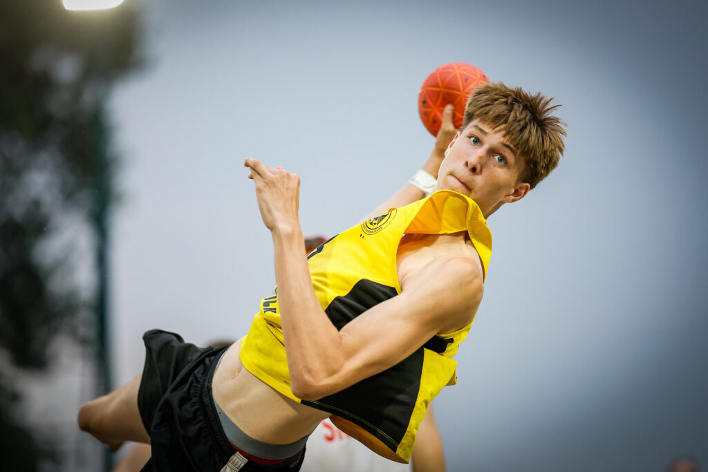 An athlete in a yellow and black uniform throws a red handball with focused intensity during a game.
