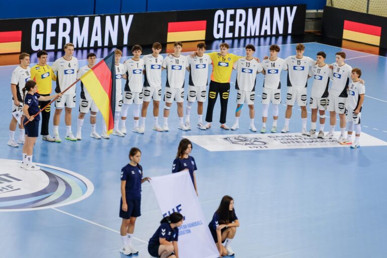 A team in white and yellow uniforms stands in a line on an indoor handball court, holding a flag, with young individuals in navy blue uniforms nearby.
