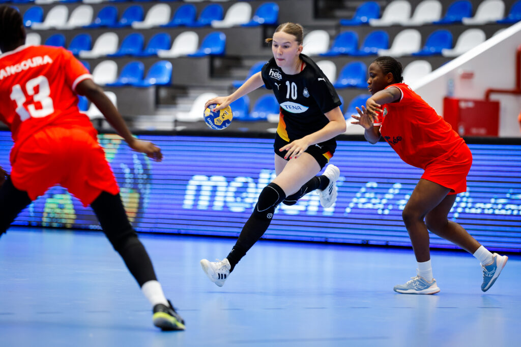 A player in a black jersey dribbles a handball while being defended by two opponents in red uniforms during an indoor game.