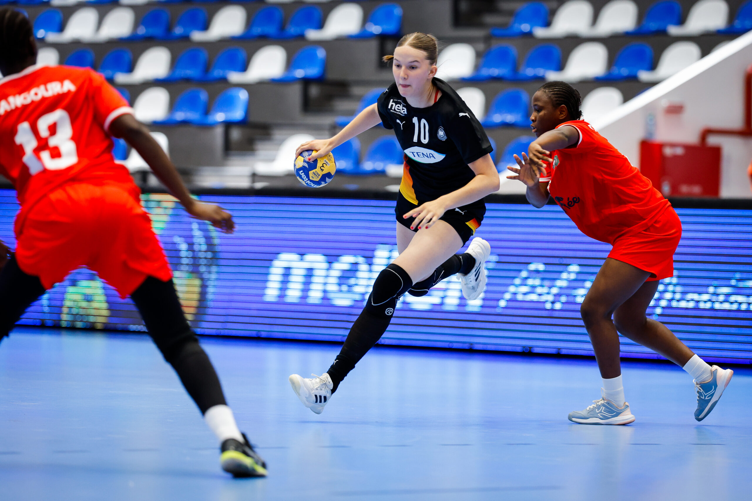 A player in a black jersey dribbles a handball while being defended by two opponents in red uniforms during an indoor game.