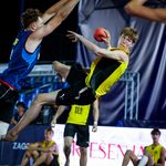 An athlete in a yellow and black uniform leaps mid-air to catch a ball while being defended by an opponent in a blue jersey during a fast-paced game.