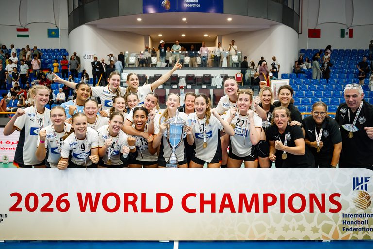 A team of athletes and staff celebrate as world champions, holding a trophy and medals in front of a banner reading "2026 WORLD CHAMPIONS."