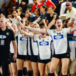 A team of athletes in white jerseys with "TENA" branding and their coach in a black shirt celebrate enthusiastically with raised arms and joyful expressions during a sports event.