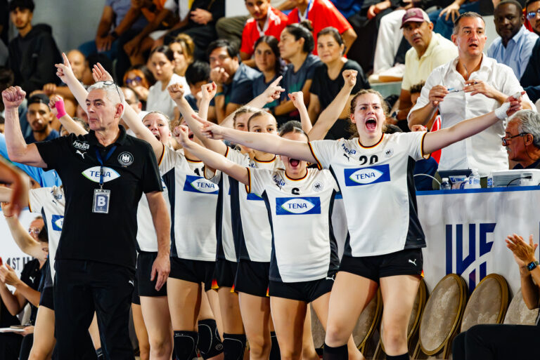 A team of athletes in white jerseys with "TENA" branding and their coach in a black shirt celebrate enthusiastically with raised arms and joyful expressions during a sports event.