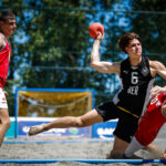 An athlete in a black jersey with the number 6 leaps to throw a ball while being challenged by an opponent on a sandy court.